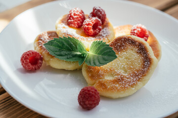 Delicious cheesecakes or pancakes with powdered sugar on white plate on wooden table with raspberries and mint leaf