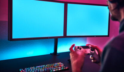 Over shoulder view of man holding game controller and playing on dual display. Blank monitor to insert your content.