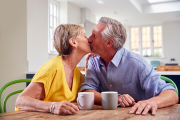 Fototapeta na wymiar Kissing Retired Couple Sitting Around Table At Home Having Morning Coffee Together