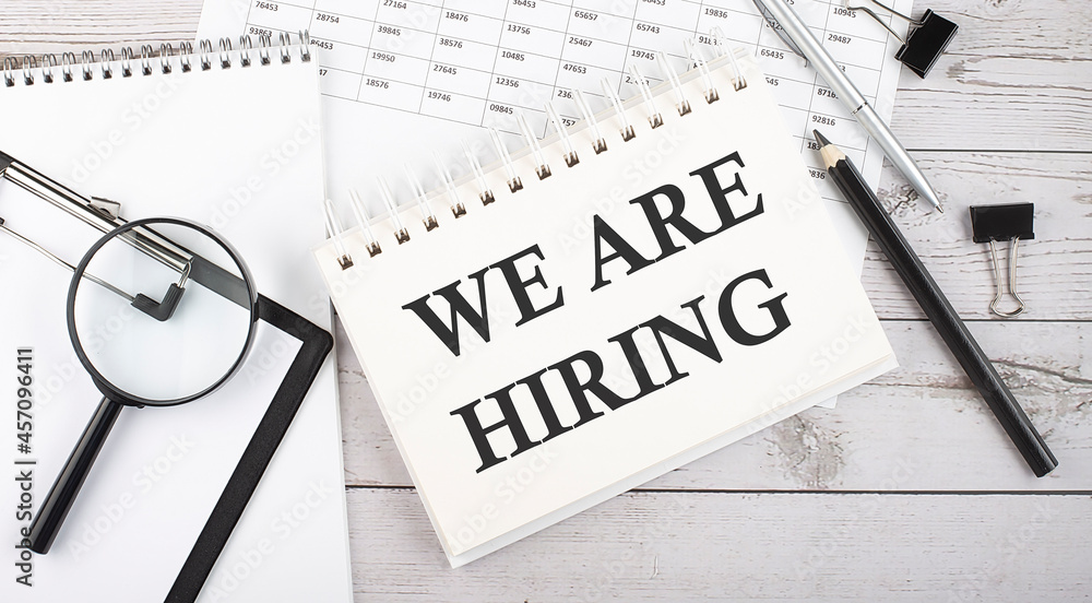 Poster notepad with text we are hiring with a pen,office tools and graphs on the desktop.