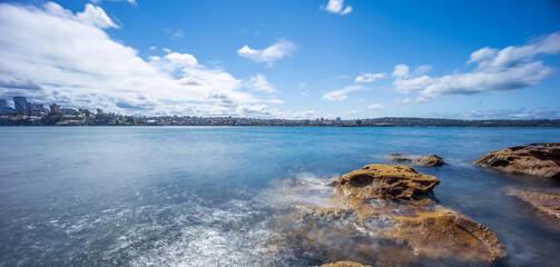 Partly cloudy Spring afternoon on Sydney Harbour with nice rocks in the foreground the soft waves crashing on the shore and the beautiful harbour foreshore as a backdrop NSW Australia