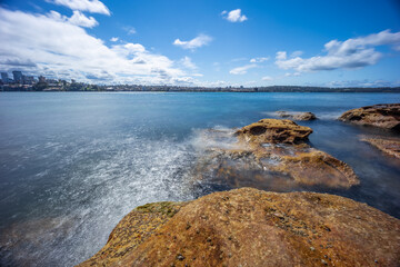 Partly cloudy Spring afternoon on Sydney Harbour with nice rocks in the foreground the soft waves crashing on the shore and the beautiful harbour foreshore as a backdrop NSW Australia