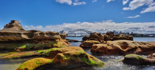 Naklejka na ściany i meble Partly cloudy Spring afternoon on Sydney Harbour with nice rocks in the foreground the soft waves crashing on the shore and the beautiful harbour foreshore as a backdrop NSW Australia