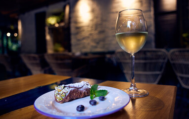 Sicilian cannoli roll with blueberries on a white ceramic plate and a glass of white wine on a restaurant table with a restaurant decor in the background.