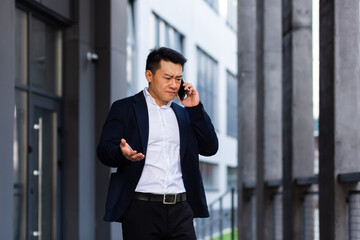 angry and serious Successful Asian businessman explains information to employees using phone, speaks near office outside