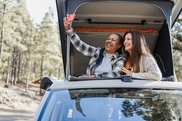 Senior women friends having fun taking selfie with mobile phone inside camper van - Focus on...