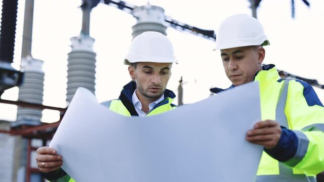 Two engineers are talking in front of electrical transmission lines, working on renewable energy development. Engineers in special clothing discuss a drawing on paper near high-voltage power line