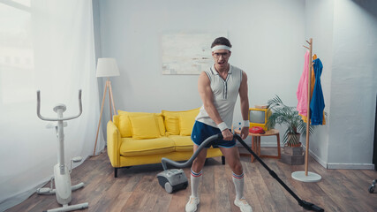 Sportsman singing and dancing while vacuum cleaning living room, retro sport concept
