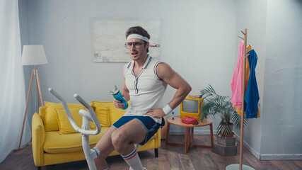 sportsman training on exercise bike and holding sports bottle in living room, retro sport concept