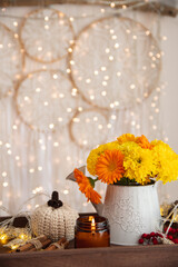 Cozy autumn atmosphere of the house. On a plaid blanket is a wooden tray with decor: a bouquet of yellow flowers, a candle, cinnamon, knitted pumpkin, garland.