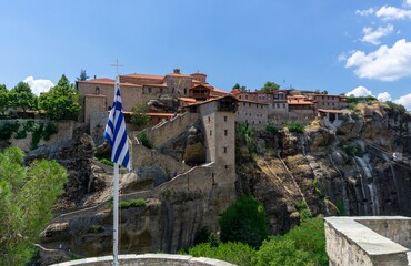 Fototapeta na wymiar View of a Meteora monastery with its stairs from the other side of the mountain, with the flag of Greece in front