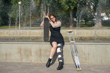 Young girl with a plastered foot in the orthosis and with crutches on a walk in the city