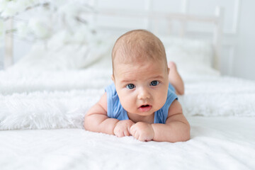 cute baby boy three months old in a blue bodysuit lies on his stomach on a white bed at home, the newborn learns to hold his head