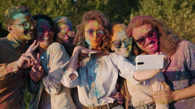 Slow motion of smiling young people dirty with bright paint taking selfie in park using smartphone camera. Holi festival and modern technology concept.
