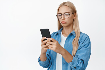 Portrait of sad and gloomy woman in glasses reading notification on mobile phone, looking at smartphone screen disappointed, white background