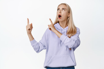 Girl in awe drop jaw, pointing fingers and looking at upper left corner, reading promo deal, showing advertisement, standing over white background