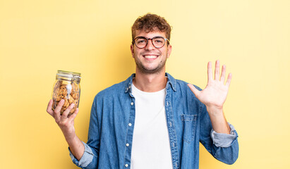 young handsome man smiling happily, waving hand, welcoming and greeting you. cookies bottle concept