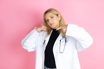 Young blonde doctor woman wearing stethoscope standing over isolated pink background doing the “call me” gesture with her hands.