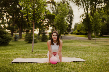 Charming fit young woman is sitting on knees on sports mat with rubber small pilates ball in park. 