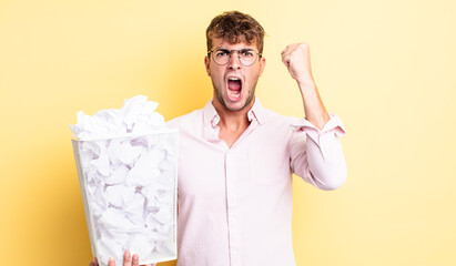 young handsome man shouting aggressively with an angry expression. paper balls trash concept