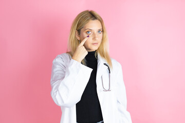 Young blonde doctor woman wearing stethoscope standing over isolated pink background Pointing to the eye watching you gesture, suspicious expression