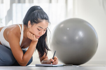 Young Asian yoga woman writing in her notebook. Sitting at home by the window after exercise. Personal growth resolutions 