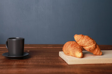 Fresh baked croissants, mug of black coffee on brown wooden table at blue wall background. Cup of espresso, bakery foods on chopping board close  up. French breakfast, morning menu, cafe concept