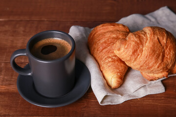 Fresh baked croissants on linen napkin, espresso cup on brown wooden table. Mug of black coffee with foam close up. Bakery food, French breakfast, morning menu, cafe concept. Top view
