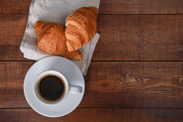 Two fresh baked croissants on linen napkin, mug of black coffee on brown wooden table. Cup of hot espresso, buns close up. Bakery food, French breakfast, morning menu, cafe concept. Top view