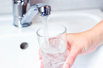 Young caucasian woman hand holding a glass with pure drinking water pouring from home faucet close up.