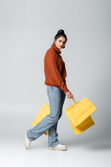 full length of brunette young woman in sweater and jeans holding shopping bags on grey