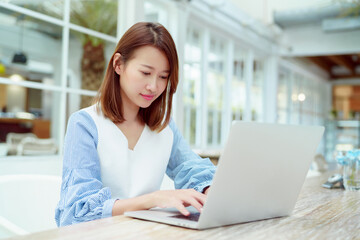 Fototapeta premium A beautiful woman wearing an Asian white shirt is sitting in front of a laptop computer shopping online with a happy smile in a bakery shop.