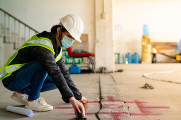 Young Asian female engineer wearing a medical mask to cover her mouth and wearing a white safety helmet using tape measure Measure the ground to mark it in the construction Zone.