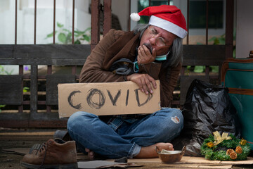 Homeless beggar wears Santa hat sitting on the floor by the street, coughing and seemingly unwell, ...