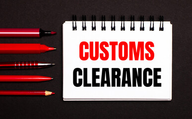 A white notebook with the text CUSTOMS CLEARANCE next to red pens, pencils and markers on a black...