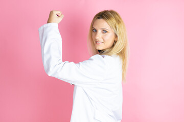 Young blonde doctor woman wearing stethoscope standing over isolated pink background feeling happy, satisfied and powerful, flexing fit and muscular biceps, looking strong after the gym