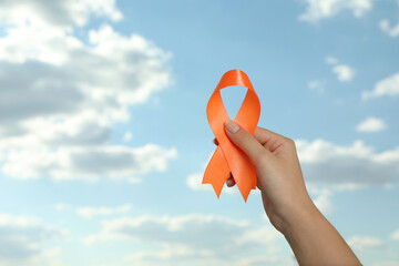 Woman holding orange ribbon against blue sky, closeup with space for text. Multiple sclerosis...