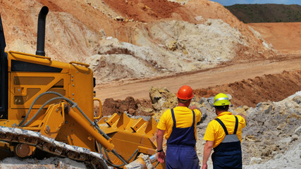 two workers monitors the extraction of clay in the quarry. Back view.  Quarry with blue clay. Big yellow excavator extracts clay. © drotik