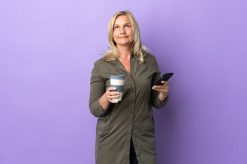 Middle age Lithuanian woman isolated on purple background holding coffee to take away and a mobile while thinking something