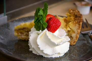 Large portion of whipped cream, decorated with strawbarry and mint, next to a piece of apple pie
