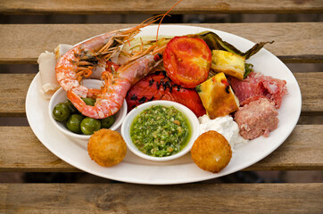 wooden table with a white plate holding a colorful array of Mediterranean snacks like shrimp, pesto, tomato and meat