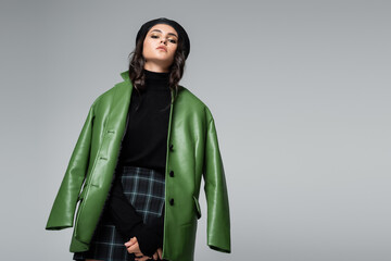 stylish woman in green leather jacket, checkered skirt and black beret posing isolated on grey