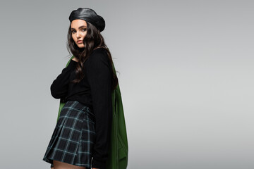 brunette woman in checkered skirt, black beret and green leather jacket posing isolated on grey