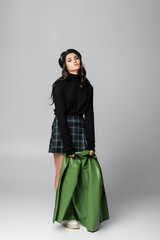 Naklejka premium full length of young stylish woman in beret and plaid skirt holding green leather jacket on grey