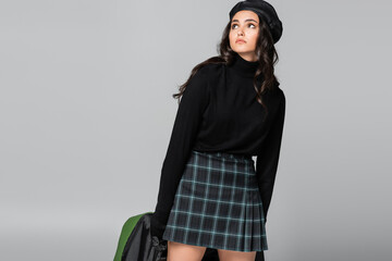 young trendy woman in beret and plaid skirt holding green leather jacket isolated on grey