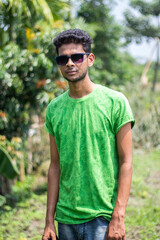 Naklejka na ściany i meble A handsome young man is standing in colorful sunglasses and a green t shirt, with a blurry green background behind him