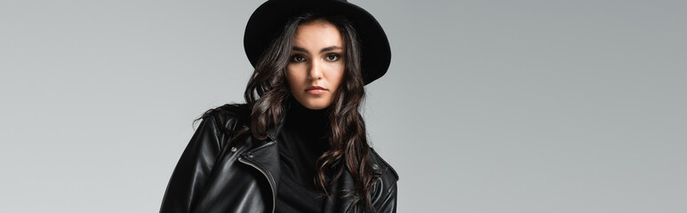 stylish woman in fedora hat and black leather jacket posing isolated on grey, banner