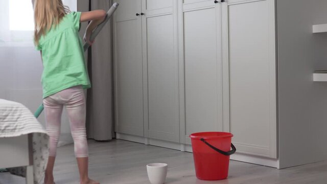 A cute little girl washes the floors in the apartment. A child with a mop is cleaned at home, the girl wets and squeezes the sex rag.