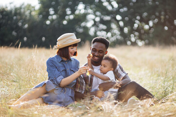 Caucasian woman and african american man sitting with their lovely son on field and eating grape. Happy family on picnic. Summer time and enjoyment concept.