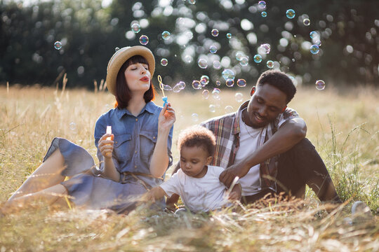 Beautiful caucasian mother and handsome african father blowing soap bubbles during summer picnic with little son on nature. Family having fun outdoors. Relaxation concept.